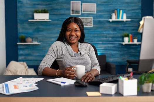 Portrait of smiling black student looking into camera while sitting at desk table in living room during home school study. African american woman browsing academic books searching information