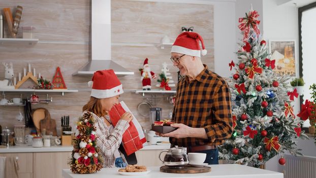 Grandchild with grandfather bringing with xmas wrapper present gift with ribbon on it celebrating christmastime during winter holiday. Happy family enjoying christmas in decorated kitchen