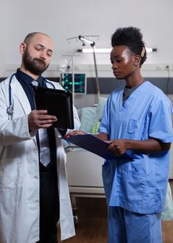 Physician doctor with asisstant with black skin checking illness recovery treatment using tablet computer during medical consultation in hospital ward. Hospitalized patient sick woman resting in bed