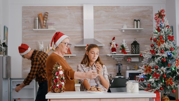 Grandmother watching xmas online video with granddaughter on tablet while grandfather bringing milk enjoying traditional festive season. Happy family celebrating christmas holiday in decorated kitchen