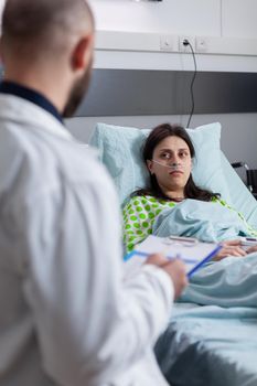 Oncologist physician doctor discussing medical expertise explaining sickness symptom to sick woman writing on clipborad. Patient sitting in bed recovering after medical surgery in hospital ward