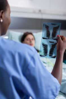 Close-up of afro american nurse analyzing lungs x-ray checking healdcare recovery. Sick woman patient resting in bed discussing disease treatment in hospital ward during therapy consultation