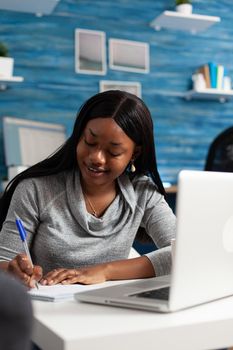 Afro american student writing math homework on notebook during high school online class using elearning platform. Black woman working remote from home studying school lesson in living room