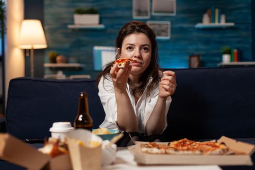 Portrait of woman watching comedy movie eating tasty delivery pizza slice relaxing on sofa in living room at night. Caucasian female enjoying takeaway food home delivered. Fastfood meal order