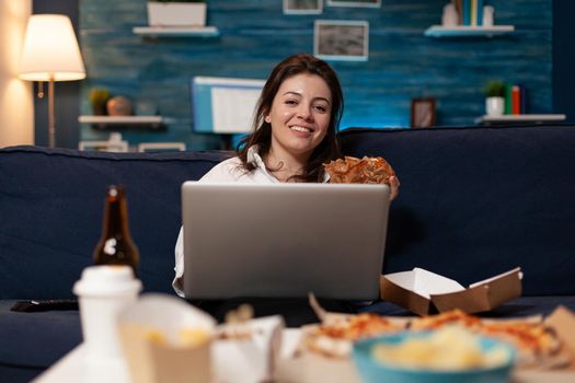 Caucasian female resting on sofa looking into camera eating tasty burger while working on laptop late at night in living room. Woman enjoying takeaway food home delivered. Fastfood meal lunch order