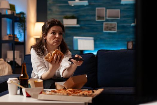 Happy woman changing channels using remote watching entertainment movie on television eating tasty delicious burger in evening. Caucasian female enjoying takeaway food home delivered