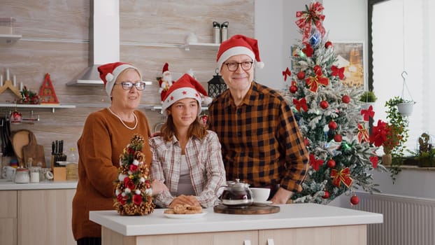 Portrait of grandparents with granddaughter spending winter holiday together standing at table in xmas decorated kitchen. Happy family wearing santa hat celebrating christmas season