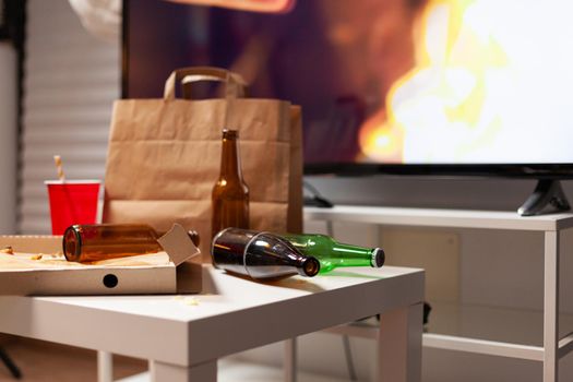 Close up of messy table with garbage food and booze leftover in empty unorganized dirty living room of depresive alone person. Untidy house apartment of woman with mental degenerating health