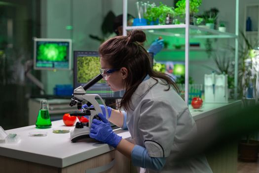 Biologist scientist in white coat working in expertise laboratory looking into microscope analyzing organic gmo leaf. Specialist researcher doing biochemistry experiment.
