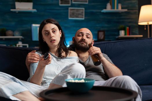 Young couple spending evening together dressed in pajamas enjoying spending time together, wife changing tv programs using remote trying to relax on couch together eating popcorn, drinking beer