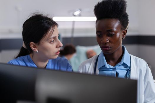 Medical team looking at computer analyzing healthcare treatment working in hospital ward. In background doctor discussing with sick man patient, recovering after illness surgery