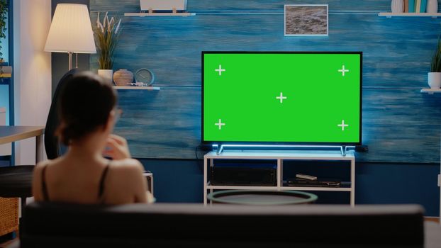 Young adult watching green screen on tv at home in modern living room. Caucasian woman having snack on couch, using mockup template display and isolated background for chroma key