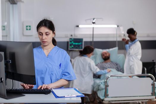 Front view of medical asisstant typing illness expertise on computer while in background physician doctors discussing with sick man. Hospitalized patient having respiratory disorder