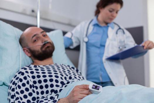 Sick man patient lying in bed with medical oximeter on finger. Specialist doctor monitoring heart pluse analyzing illness cardiogram working in hospital ward explaining disease diagnosis