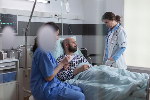 Woman nurse writing sickness diagnosis on clipboard while specialist doctor in uniform monitoring disease symptom analyzing medical recovery in hospital ward. Sick man patient resting in bed