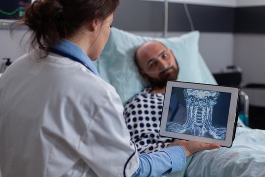 Physician practitioner doctor analyzing bones radiography using tablet computer in hospital ward. Hospitalized man patient with nasal oxygen tube resting in bed during healthcare recovery