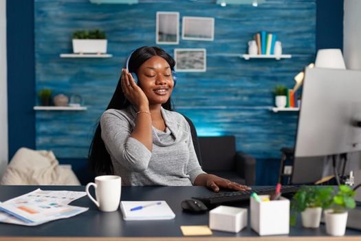 African american student wearing headphones listening happy music sitting at desk in living room. Woman with black skin working remote from home browsing academic courses using elearning platform