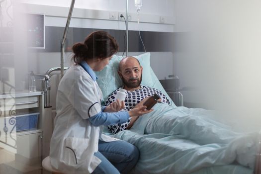 Sick man patient with nasal oxygen tube resting in bed during medical examination in hospital ward. Specialist woman doctor explaining pill treatment against pain discussing pharmacy prescription