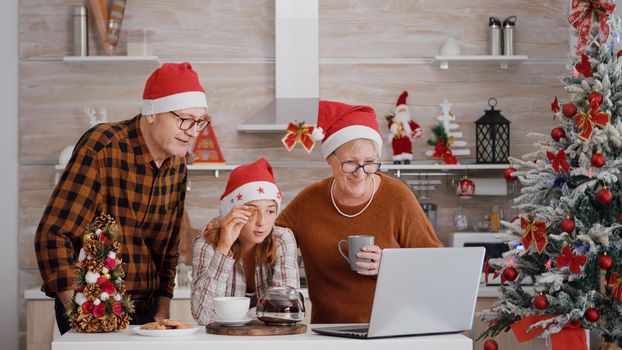 Granddaughter with grandparents discussing with remote parents during online videocall meeting on laptop while eating baked cookies in xmas decorated kitchen. Family enjoying spending time together