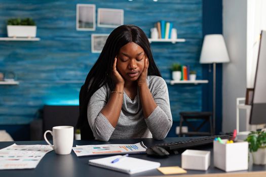 Tired unhappy frustration african student having headache pain sitting at desk in living room. Worried sick upset black american woman suffering forehead migraine working remote from home