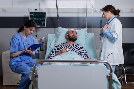 Patient talking with medical doctors while sitting in bed during disease recovery in hospital ward. Practitioner writing sickness treatment on clipboard checking heart pulse