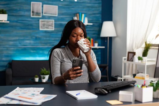 Black woman holding smartphone in hands chatting with people browsing communication information sitting at desk in living room. African american student looking on social media