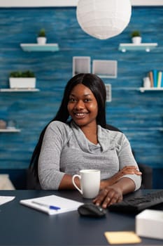 Portrait of african american student sitting at desk table in living room looking into camera smiling during homeschooling. Black adolescent working remote at high school information from home