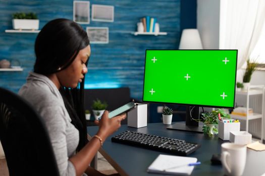 African american woman browsing school information on phone working remote from home in living room. On table standing mock up green screen chroma key computer with isolated display