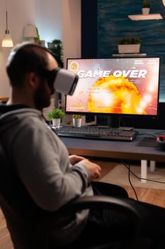 Professional player wearing vr goggles sitting on gaming chair playing online shooter game. Defeated man losing video game tournament using joystick late at night in living room.