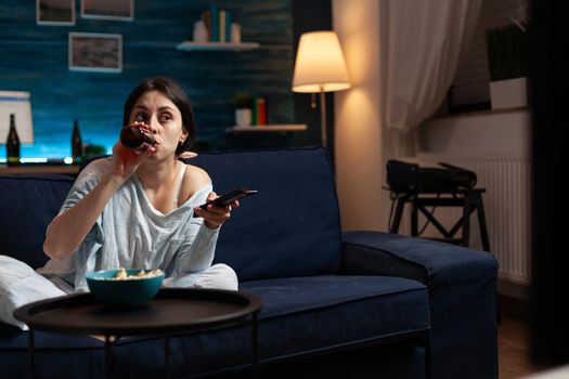 Confused young woman drinking beer while watching movie on tv changing channels using remote alone in living room. Female feeling confused having shocked facial expression relaxing at night