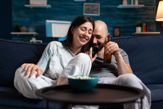 Relaxed couple in pajamas relaxing on sofa eating popcorn watching comedy movie, enjoying free time together. Husband and wife relaxing late at night in living room. Looking at entertainment film