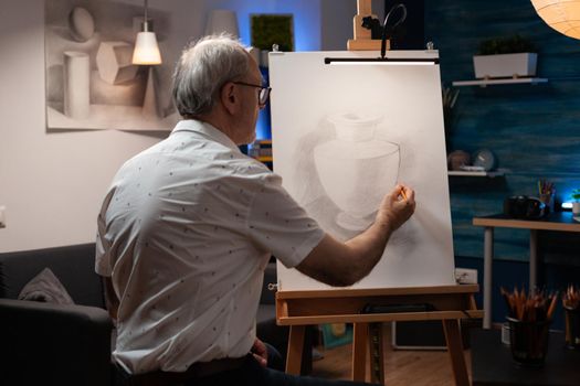 Elderly artist working on vase drawing with pencil on canvas and easel at art studio. Caucasian senior man recreating authentic masterpiece with object for fine art project. Old creative person