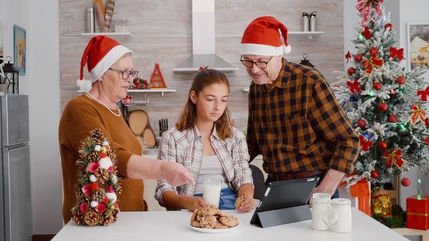 Happy family watching online xmas movie on tablet computer enjoying holiday season standing at table in decorated kitchen. Grandchild eating baked cookies celebrating christmas tradition