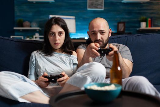 Focused determined gamers playing soccer videogame late at night trying to win online competition. Couple sitting on sofa, gaming video games together using joystick, wireless controller