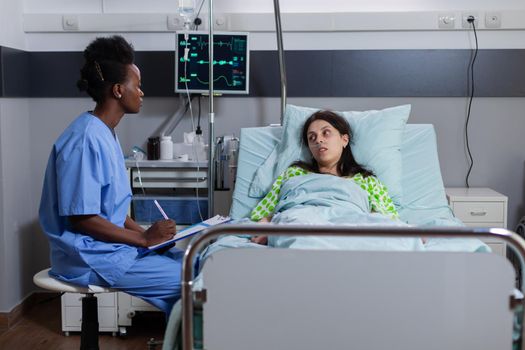 Afro american asisstant monitoring sick woman discussing sickness symptom writing medical expertise on clipboard during recovery appointment. Patient resting in bed in hospital ward