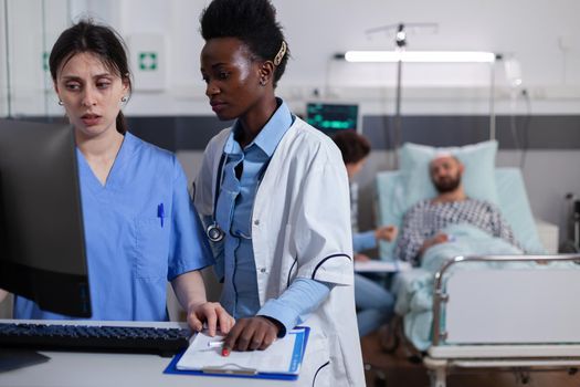 Woman nurse explaining medical recovery on computer to afro american practitioner in hospital ward. Hospitalized man patient discussing with doctor illness treatment during expertise consultation