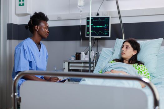 Sick woman with nasal oxygen tube explaining disease symptom during medical consultation in hospital ward. Black asisstant monitoring heartpulse writing sickness treatment on clipboard