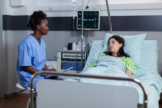 Sick woman with nasal oxygen tube resting in bed during medical consultation discussing with specialist doctor disease symptom in hopistal ward. Black practitioner writing sickness treatment