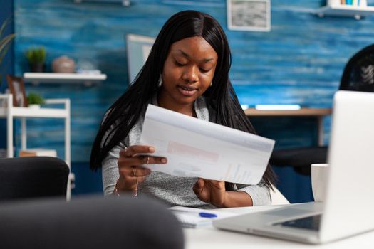 Afro american woman analyzing financial graph document remote working at marketing presentatrion in living room. Smart black student checking accounting infographics planning statistics at home