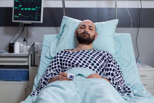 Sick man looking into camera while sitting in bed waiting for disease treatment recovering after respiratory surgery in hospital ward. Hospitalized patient wearing nasal oxygen tube