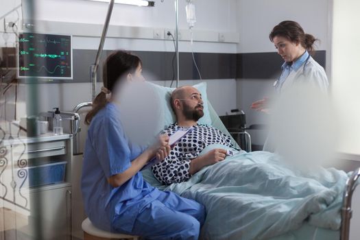 Patient talking with doctors while resting in bed during disease recovery in hospital ward. Practitioner woman asisstant writing sickness treatment on clipboard examining medical symptom