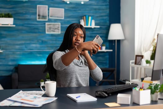 Blogger black student smiling while taking selfie with smartphone sitting at desk table in living room. Attractive african woman browsing sharing lifestyle photo on social media app