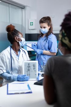 African american doctor with face mask against coronavirus discussing bones radiography with nurse during clinical appointment in hospital office. Radiologist asisstant writing medication treatment