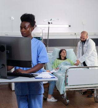 Black nurse typing disease symptom while specialist practitioner doctors monitoring sick woman checking medical expertise during rehabilitation appointment. Patient resting in bed in hospital ward