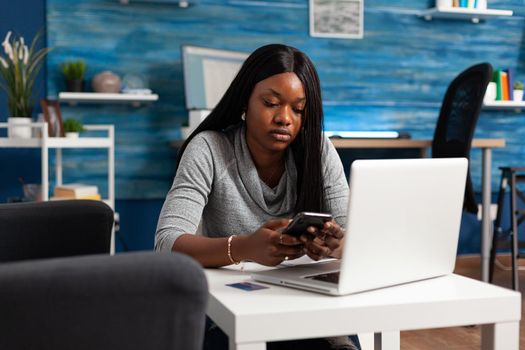 African american student holding phone in hands texting message chatting with group friends working remote from home. Black woman browsing on internet looking on social media in living room