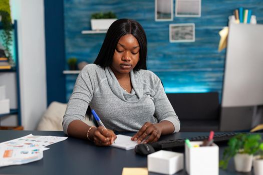 African american student writing high school homework on notebook during online communication course working remote from home. Black woman studying business ideas using elearning platform