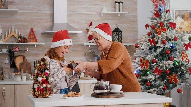 Grandmother bringing wrapper present gift with ribbon on it to granddaughter enjoying christmastime in xmas decorated kitchen. Happy family wearing santa hat celebrating christmas holiday