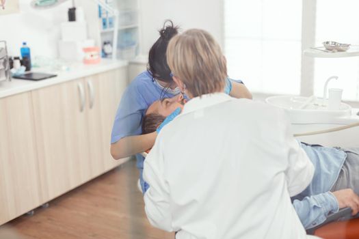 Sick patient sitting on dental chair with open mouth during stomatology procedure. Senior woman stomatologist and hospital nurse with masks holding sterilized tools checking teeth health