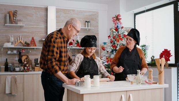 Grandchild wearing cooking apron preparing homemade delicious cookies dough using rolling pin while grandfather bringing gingerbread ingredients. Happy family celebrating christmas holidays