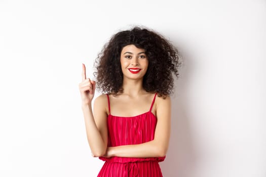 Beautiful smiling lady in red dress showing number one, raising finger and looking pleased, standing over white background.